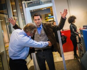Ein junger Mann steht mit erhobenen Armen in der Sicherheitsschleuse eines Flughafens. Ein Sicherheitsmitarbeiter in einem hellblauen Hemd tastet ihn im Bereich der Taille ab. Im Hintergrund sind ein Metalldetektorrahmen, eine weitere Person an der Gepäckkontrolle sowie ein Gepäckscanner zu sehen.